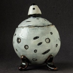May include: A small, white ceramic jar with a lid and three legs. The jar is decorated with black and grey dots and swirls.