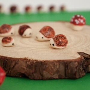 Peut inclure: Une collection de petits hérissons en céramique brun et blanc sur une tranche de bois. Les hérissons sont disposés en cercle, avec un au centre. Il y a un champignon rouge sur la tranche de bois.