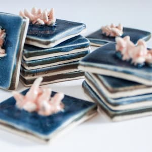 May include: A collection of square ceramic tiles in varying stacks. Each tile is a deep blue with a textured surface and a raised coral-like design in a soft pink hue. The tiles are arranged on a white surface.