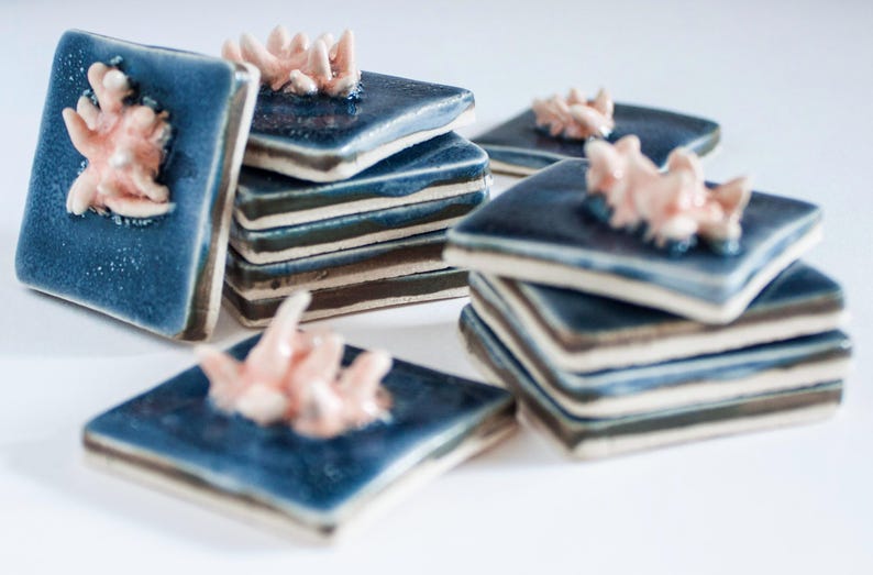 May include: A collection of square ceramic tiles in varying stacks. Each tile is a deep blue with a textured surface and a raised coral-like design in a soft pink hue. The tiles are arranged on a white surface.