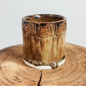May include: A handcrafted ceramic tumbler with a distinctive design. The cup has a brown and tan glaze, a textured surface, and a white base. The top rim features a repeating square spiral pattern.