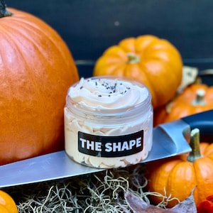 May include: A seasonal display featuring orange pumpkins and gourds, with autumn leaves. A jar of cream, labelled "THE SHAPE", rests on a large knife. The background is a dark wooden surface.