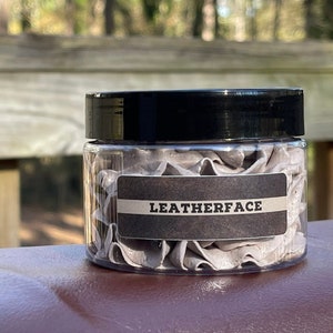 May include: A clear plastic jar with a black lid, filled with gray, folded material. A dark brown label with the word "LEATHERFACE" in white is affixed to the jar. The jar is set on a brown surface, with a blurred outdoor background.