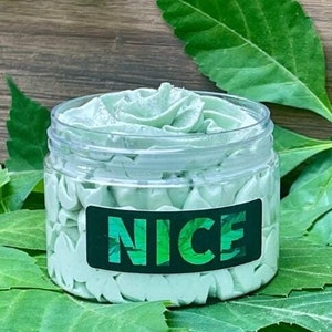 Puede incluir: Un frasco transparente lleno de manteca corporal batida de color verde claro, con la palabra "NICE" en verde y blanco en una etiqueta. El frasco está rodeado de hojas verdes, con un fondo de madera.