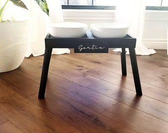 May include: A black elevated pet feeder with two white ceramic bowls. The feeder has a rectangular top and four angled legs. The name "Gertie" is written in script on the front. The feeder is on a hardwood floor.