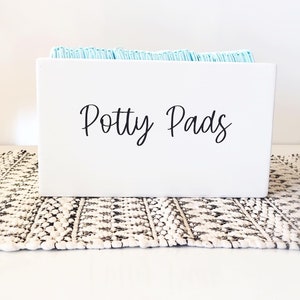 May include: White rectangular box with the words "Potty Pads" written on the front. The box is filled with blue potty pads.