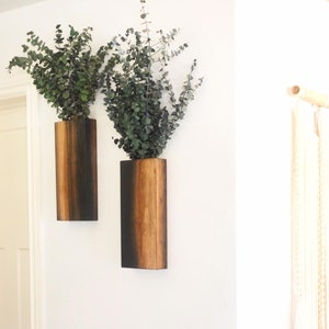 May include: Two wooden wall vases with a dark brown stain and a light brown wood grain. The vases are mounted on a white wall and each holds a bunch of green eucalyptus.