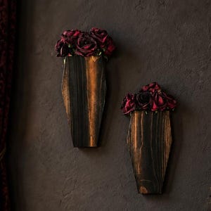 May include: Two wall-mounted wooden coffin-shaped planters featuring dark wood grain finishes, each filled with deep red artificial roses. These decorative wall vessels are positioned vertically on a dark textured wall above a black velvet sofa.