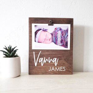 May include: A brown wooden photo frame with a clip to hold a photo. The frame has the name "Vanna James" written in white on it. A photo of a baby is in the frame.