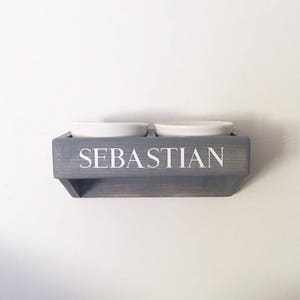 Puede incluir: Un soporte de cuenco para comida de mascotas de madera gris con el nombre "Sebastian" en la parte delantera. Dos cuencos de cer&aacute;mica blanca est&aacute;n montados en el soporte.