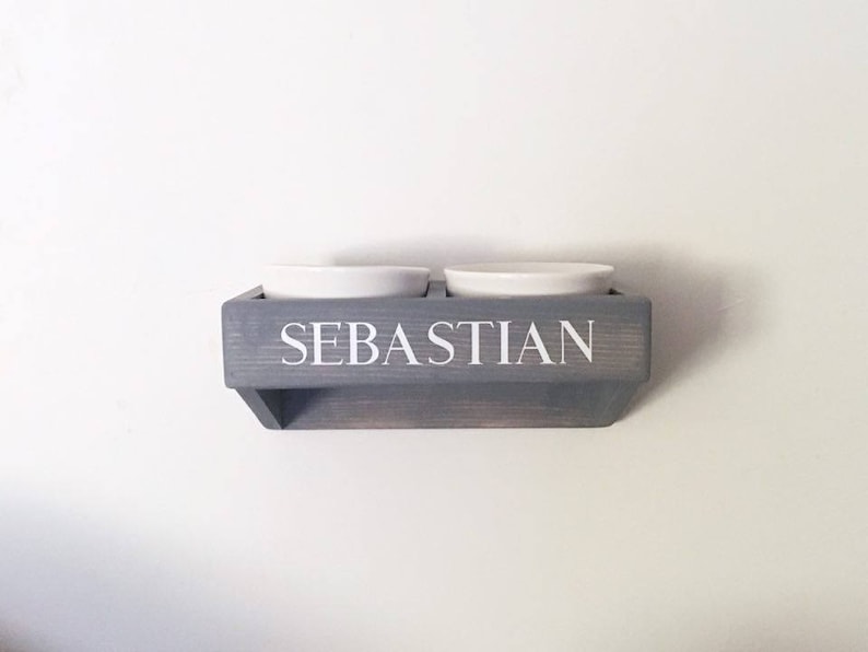 Puede incluir: Un soporte de cuenco para comida de mascotas de madera gris con el nombre "Sebastian" en la parte delantera. Dos cuencos de cer&aacute;mica blanca est&aacute;n montados en el soporte.