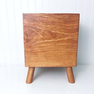 May include: A wooden storage box with a light brown stain. The box has a square shape and sits on four short, tapered legs.