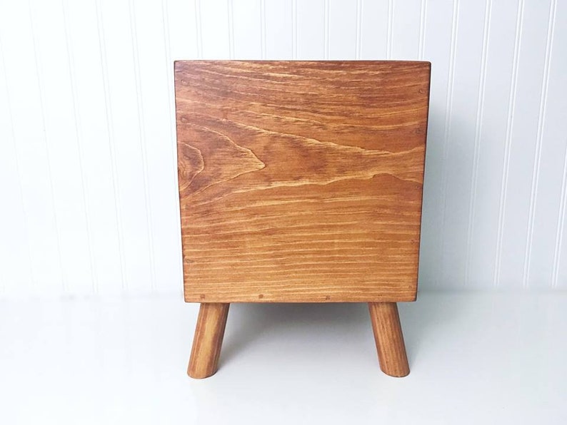May include: A wooden storage box with a light brown stain. The box has a square shape and sits on four short, tapered legs.