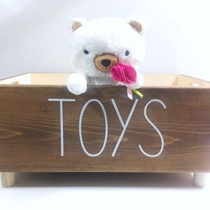 May include: A wooden toy storage box with a white label that says "TOYS". The box has a white teddy bear with a pink flower in its mouth peeking out of the top.