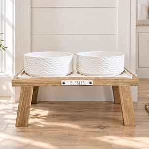May include: A raised wooden pet feeder stand with four angled legs, featuring two white ceramic bowls with a textured scalloped pattern. A small rectangular metal nameplate is attached to the front center, clearly displaying the name BUBBLES.