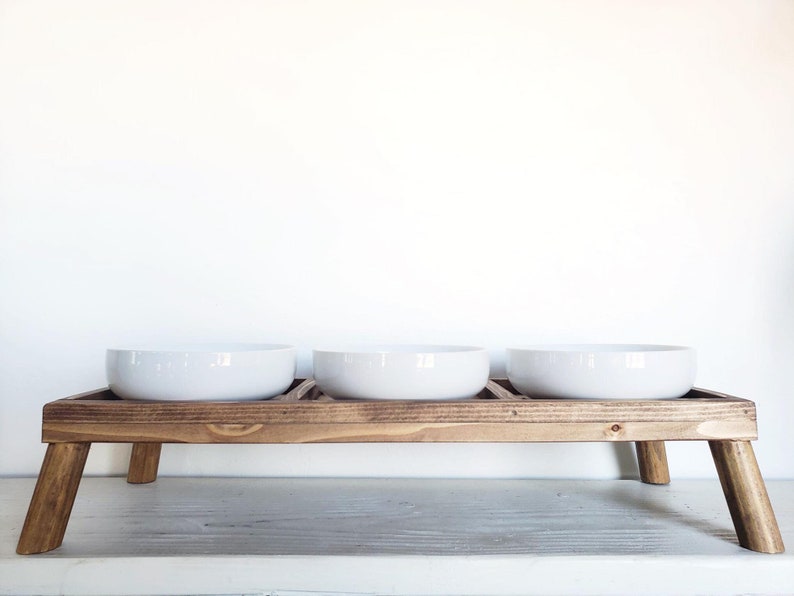 Puede incluir: Un soporte de madera para comida de mascotas con tres cuencos de cer&aacute;mica blanca. El soporte tiene cuatro patas y est&aacute; hecho de madera marr&oacute;n.