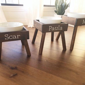 May include: Three elevated pet feeding stations made of dark stained wood, each featuring four angled legs and a white ceramic bowl. The front of each wooden frame is personalized with white lettering reading Scar, Paula, and Max respectively.