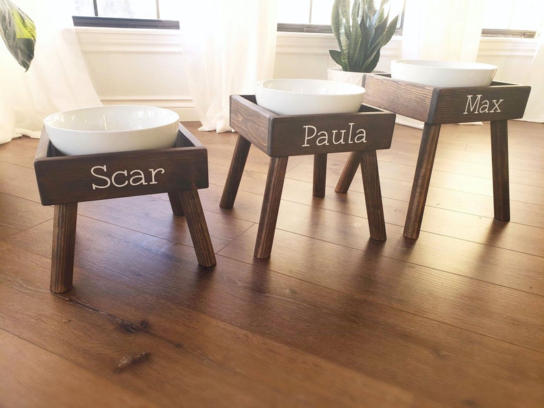 May include: Three elevated pet feeding stations made of dark stained wood, each featuring four angled legs and a white ceramic bowl. The front of each wooden frame is personalized with white lettering reading Scar, Paula, and Max respectively.