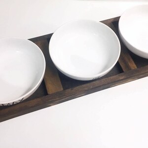 May include: Three white ceramic bowls with black lettering on a wooden tray. The bowls are arranged in a row on the tray, which is made of dark brown wood.