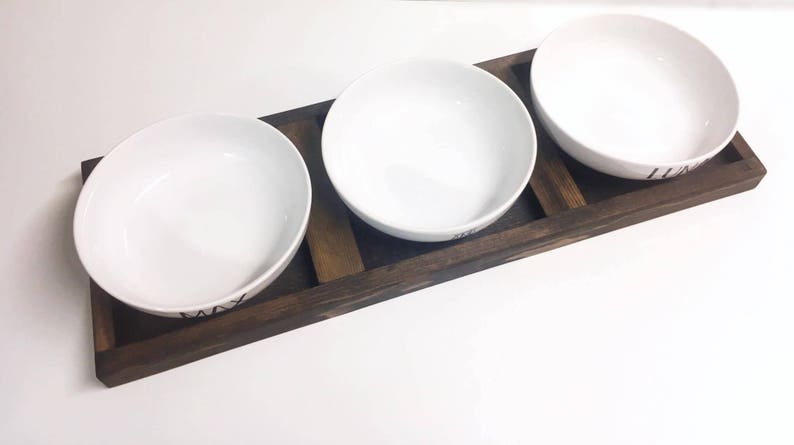 May include: Three white ceramic bowls with black lettering on a wooden tray. The bowls are arranged in a row on the tray, which is made of dark brown wood.