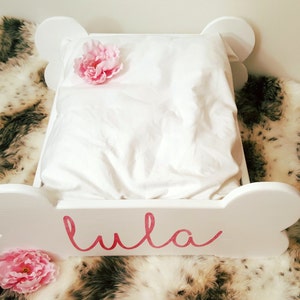 May include: A white dog bed in the shape of a bone, featuring a white pillow and a pink flower. The name "Lula" is written in pink script on the front. The bed rests on a faux fur blanket.