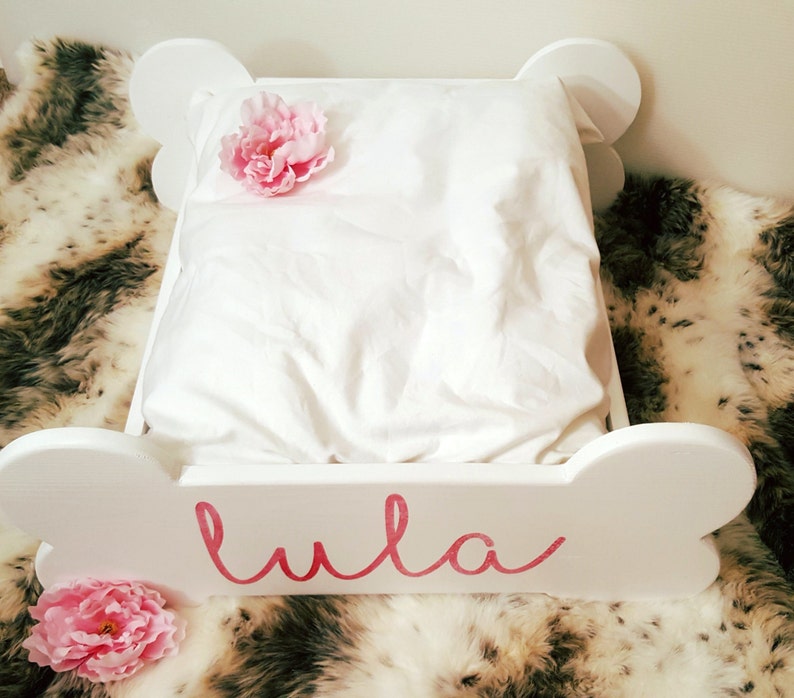 May include: A white dog bed in the shape of a bone, featuring a white pillow and a pink flower. The name "Lula" is written in pink script on the front. The bed rests on a faux fur blanket.