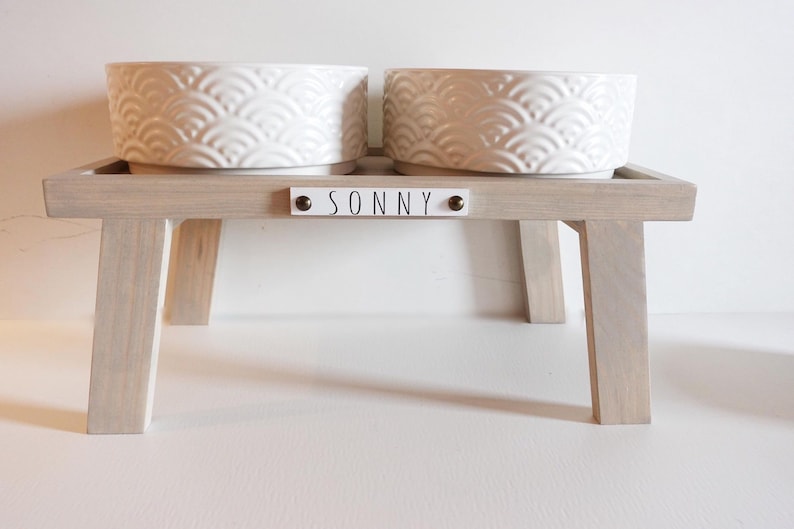 May include: White ceramic pet bowls with a raised design on a light grey wooden stand with the name "SONNY" on a white plaque.