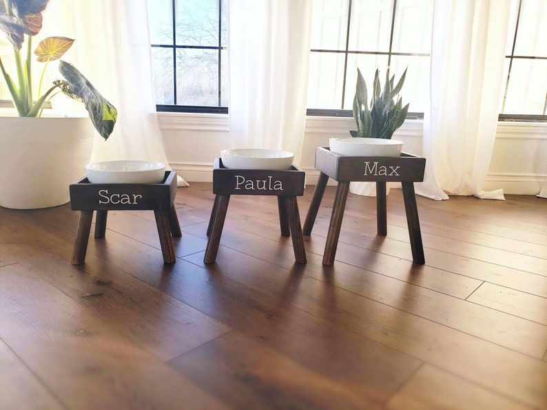May include: Three dark brown wooden pet feeding stands of varying heights, each featuring a white ceramic bowl on top. The front of each stand is personalized with white lettering reading Scar, Paula, and Max respectively, with four legs.
