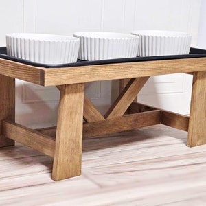 May include: A wooden pet food stand with a black tray and three white ceramic bowls. The stand has a rustic brown finish and a simple design.