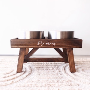 May include: A wooden dog bowl stand with two stainless steel bowls. The stand is stained a dark brown and has the name "Barley" engraved on the front.