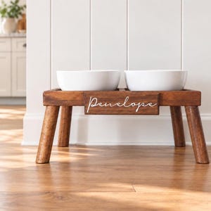 May include: A dark brown wooden elevated pet feeder stand with four sturdy legs and two white ceramic bowls. The front of the stand features a rectangular wooden plaque with the name Penelope written in white cursive script lettering.
