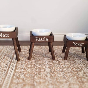 May include: Three elevated pet feeding stations with dark brown wooden stands and white ceramic bowls. Each stand features four tapered legs and a front panel with white lettering reading Max, Paula, and Scar, arranged in descending height order.