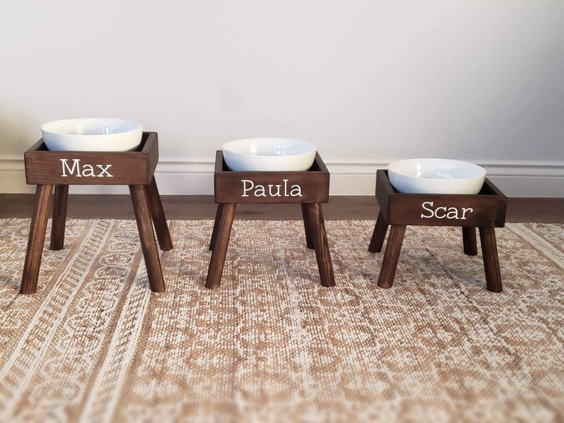 May include: Three elevated pet feeding stations with dark brown wooden stands and white ceramic bowls. Each stand features four tapered legs and a front panel with white lettering reading Max, Paula, and Scar, arranged in descending height order.