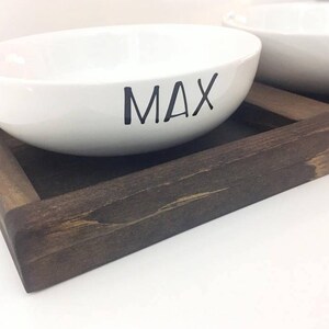 May include: Two white ceramic pet bowls with the name "MAX" written on one bowl, sitting on a dark brown wooden tray.