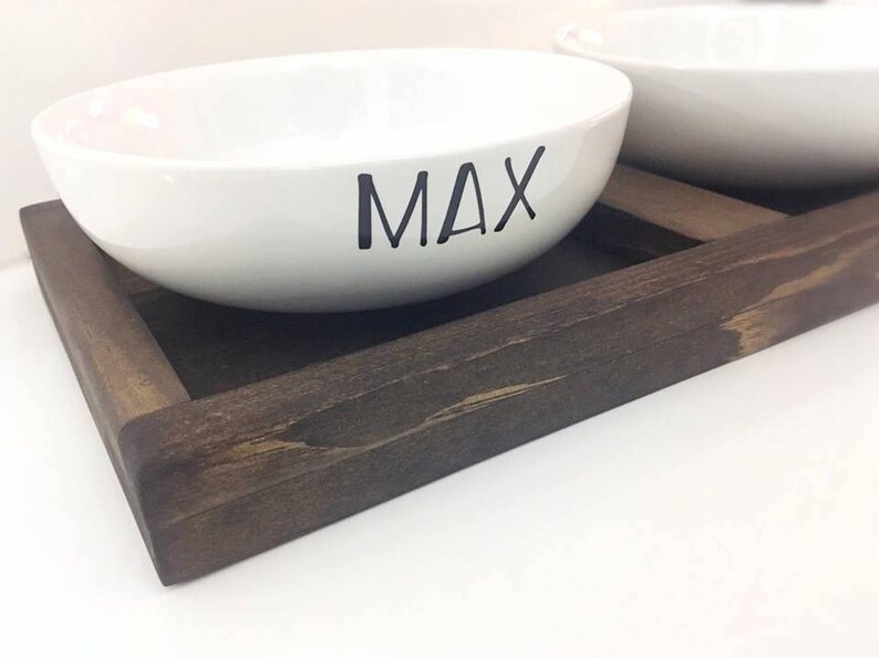 May include: Two white ceramic pet bowls with the name "MAX" written on one bowl, sitting on a dark brown wooden tray.