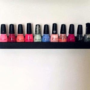 May include: A black shelf with 10 bottles of nail polish in various colors, including pink, red, blue, and gray. The shelf is mounted on a white wall.