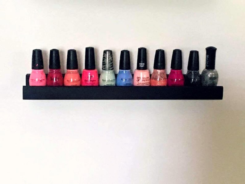 May include: A black shelf with 10 bottles of nail polish in various colors, including pink, red, blue, and gray. The shelf is mounted on a white wall.