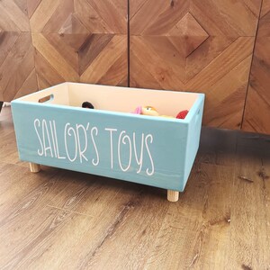 May include: A light blue wooden toy box with the words "Sailor's Toys" written in white on the front. The box has four small wooden legs and is filled with toys.