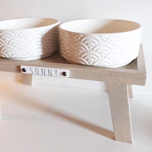 May include: White ceramic pet bowls with a raised design on a wooden stand with the name "SONNY" on the front.
