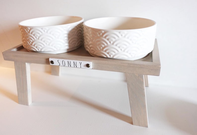 May include: White ceramic pet bowls with a raised design on a wooden stand with the name "SONNY" on the front.