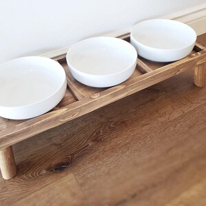 Puede incluir: Un soporte de madera para comida de mascotas con tres cuencos de cer&aacute;mica blanca. El soporte tiene dos patas y est&aacute; te&ntilde;ido de un color marr&oacute;n oscuro.