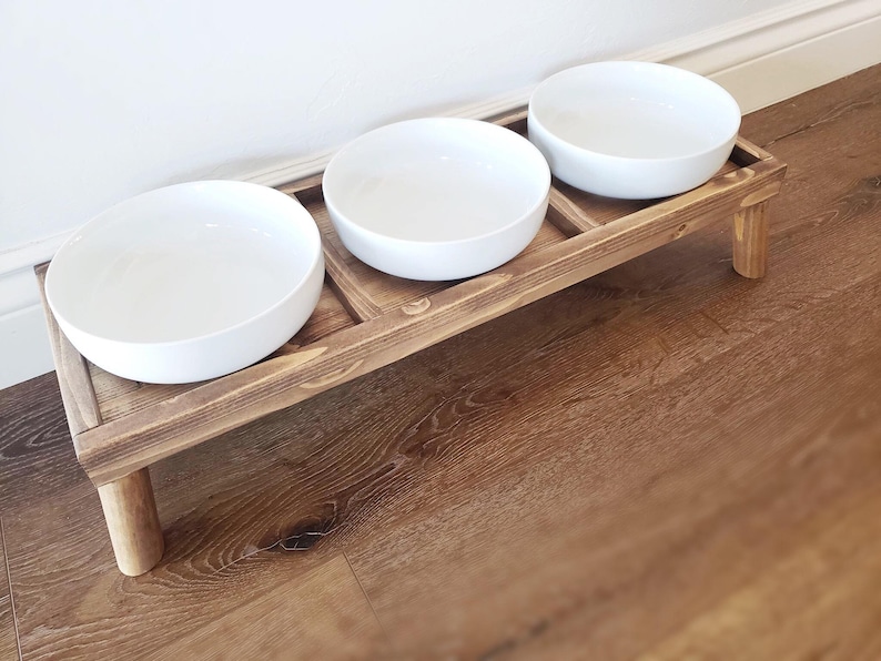 Puede incluir: Un soporte de madera para comida de mascotas con tres cuencos de cer&aacute;mica blanca. El soporte tiene dos patas y est&aacute; te&ntilde;ido de un color marr&oacute;n oscuro.