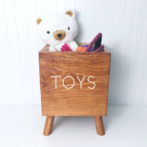 May include: A wooden toy storage box with four legs. The box is stained a dark brown color and has the word "TOYS" written on the front in white paint. The box is filled with toys, including a white stuffed bear and a purple rope toy.