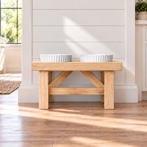 May include: A light brown wooden pet feeder stand with a rectangular top and four sturdy legs connected by diagonal support braces. Two white ceramic bowls with a fluted, ribbed exterior design are placed side by side on the wooden surface.