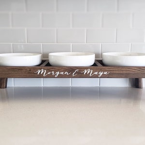 Puede incluir: Un soporte de madera para comida de mascotas con tres cuencos de cer&aacute;mica blanca. El soporte tiene cuatro patas y est&aacute; grabado con los nombres "Morgan & Maya".