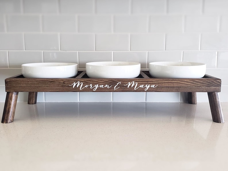 Puede incluir: Un soporte de madera para comida de mascotas con tres cuencos de cer&aacute;mica blanca. El soporte tiene cuatro patas y est&aacute; grabado con los nombres "Morgan & Maya".
