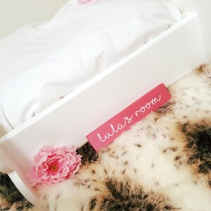 May include: A white wooden cradle with a white pillow and bedding, set on a faux fur blanket. A pink sign with the text "Lula's room" is placed on the cradle. Pink flowers are placed near the cradle.