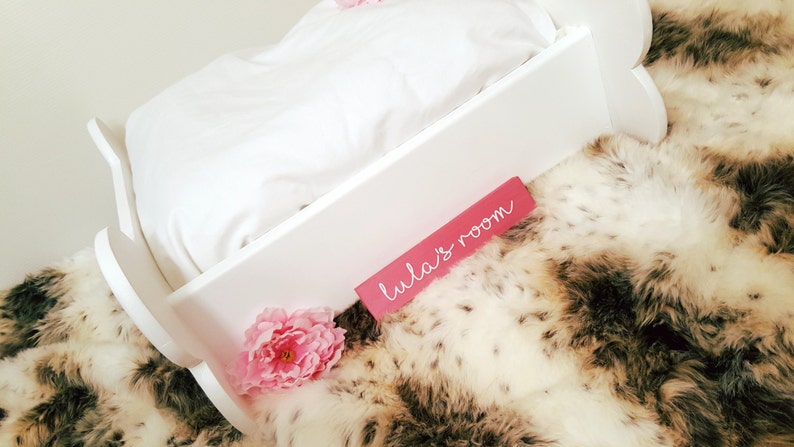 May include: A white wooden cradle with a white pillow and bedding, set on a faux fur blanket. A pink sign with the text "Lula's room" is placed on the cradle. Pink flowers are placed near the cradle.