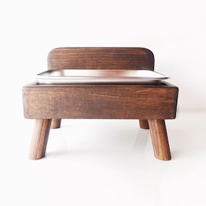 May include: A wooden pet food bowl stand with a raised edge and four legs. The bowl is made of stainless steel.