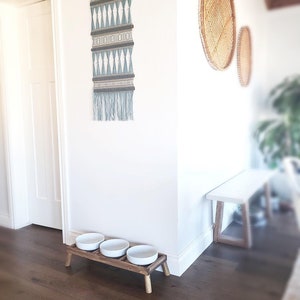 Puede incluir: Un soporte de madera para comida de mascotas con tres cuencos de cer&aacute;mica blanca. El soporte tiene cuatro patas y est&aacute; en una habitaci&oacute;n con suelo de madera dura. Hay un tapiz de pared tejido y una cesta tejida en la pared detr&aacute;s del soporte.